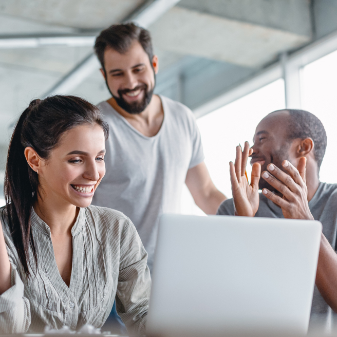 Mulher sorrindo para o computador e dois homem, um branco e preto ajudando, sendo o preto está aplaudindo-a